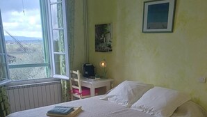 Cama bem arrumada com lençóis brancos e toalhas dobradas, uma mesinha com abajur e um quadro na parede.