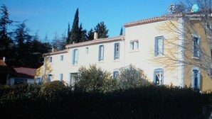 Um edifício de dois andares com fachada clara e telhado de telhas vermelhas. Possui várias janelas com persianas e é cercado por um pequeno muro de sebe.