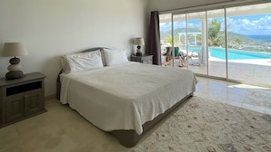 Un dormitorio amplio con una cama grande, una mesita de noche, una lámpara y vistas a una piscina y montañas a través de amplios ventanales.