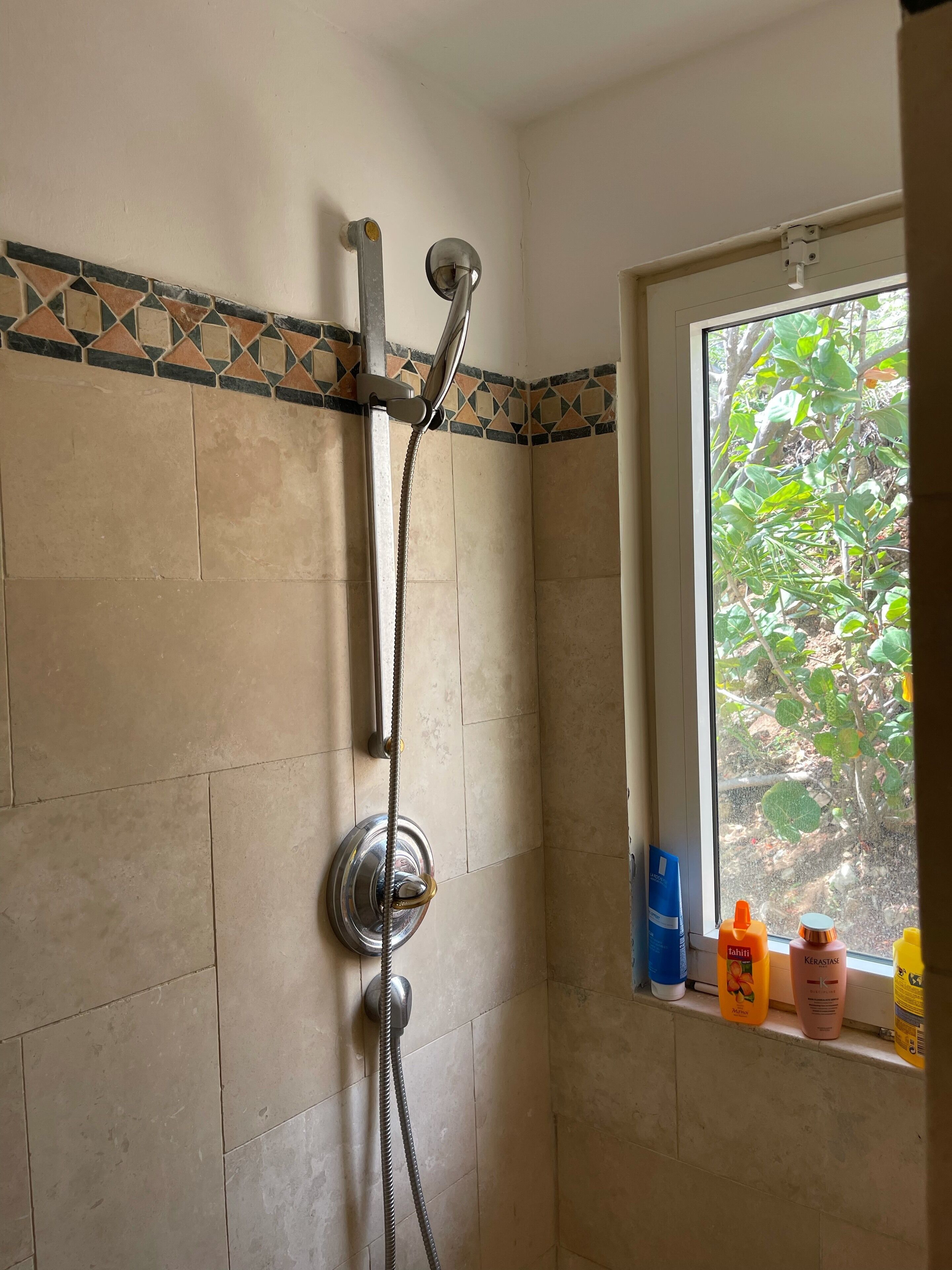 A bathroom with a shower, tiled walls, a window with a view of greenery, and toiletries on the windowsill.