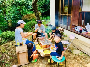 Um grupo de pessoas reunido em volta de uma mesa com comida e bebida, ao ar livre, com vegetação ao fundo.