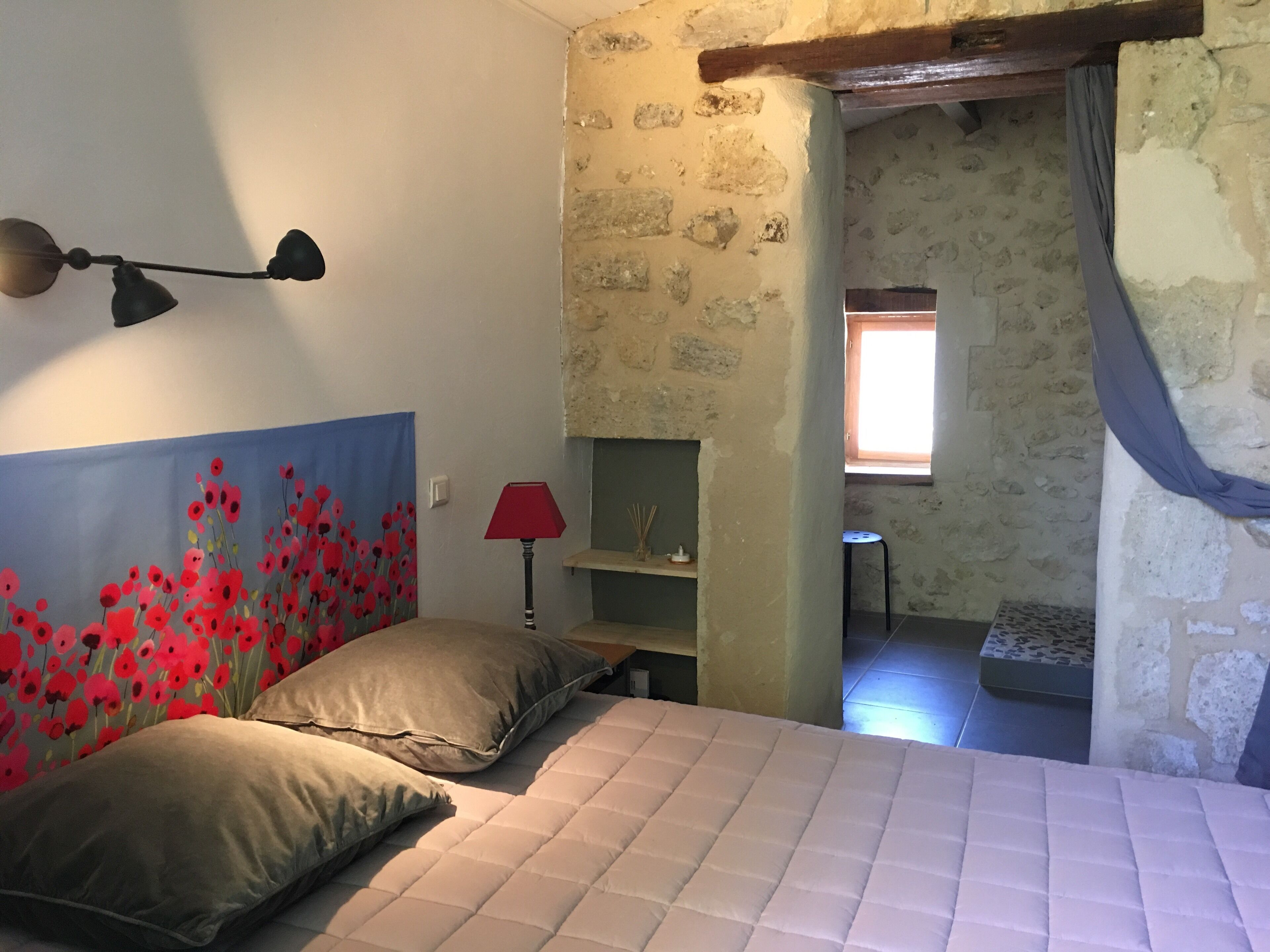 Une chambre avec un lit, une lampe fixée au mur, une lampe rouge sur une table de chevet, un tableau représentant des fleurs rouges, un mur en pierre, une fenêtre avec des rideaux et un petit tabouret.