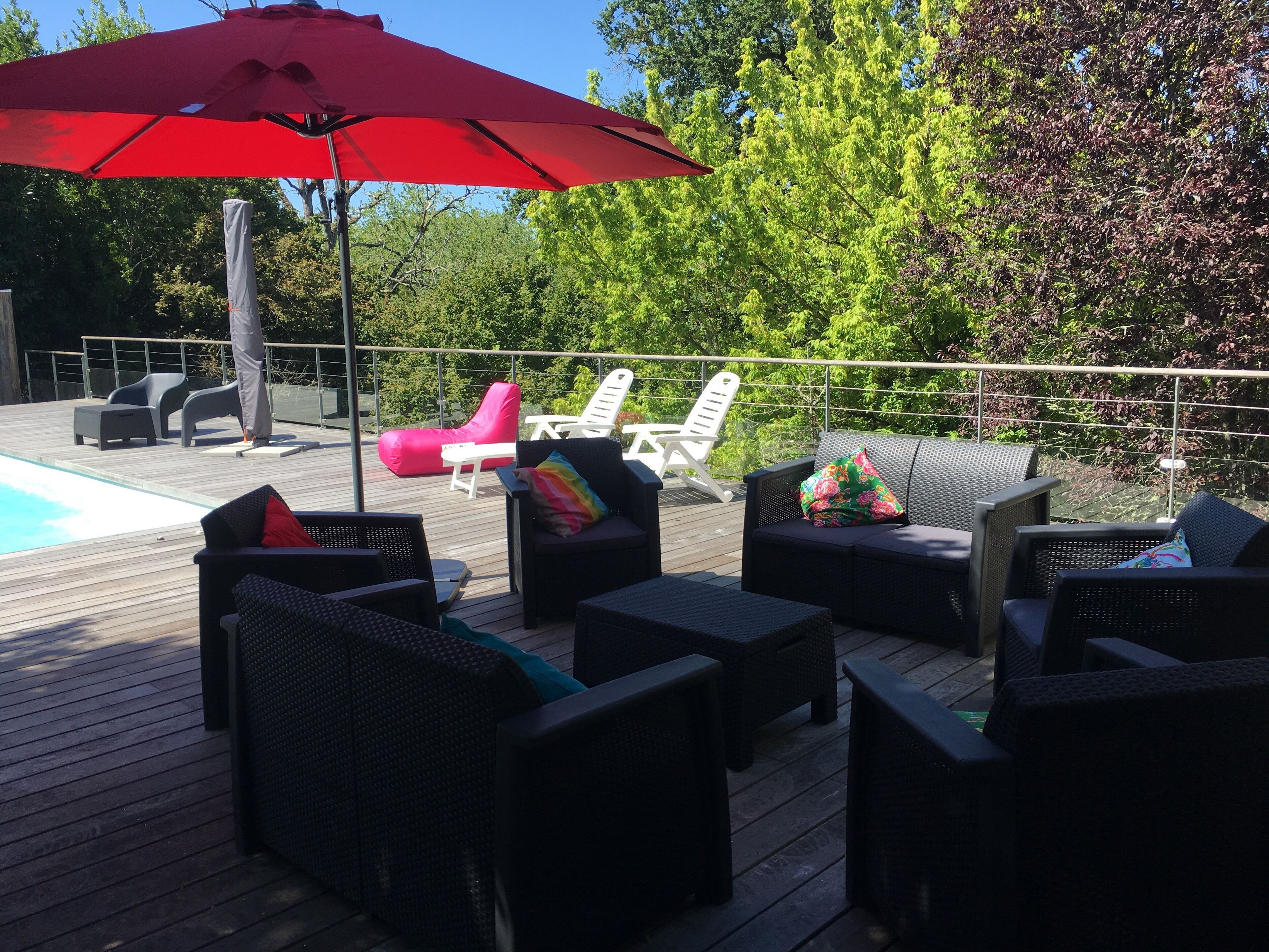 Een terras buiten met rieten meubilair, een rode parasol en een zwembad op de achtergrond.