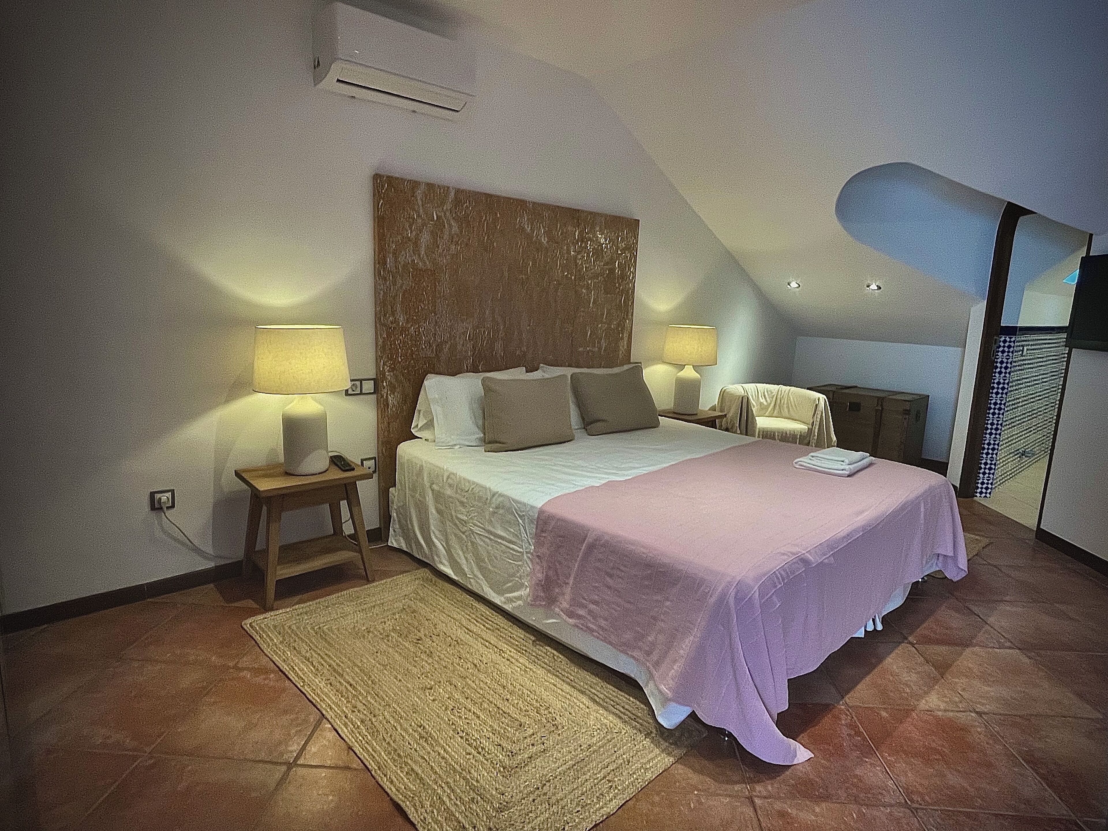 A bedroom with a large bed, two bedside tables with lamps, a wooden headboard, a small rug, and a view of the outside through a window.