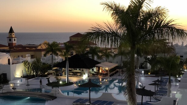 A resort with a pool, lounge chairs, and a view of the sea at sunset.
