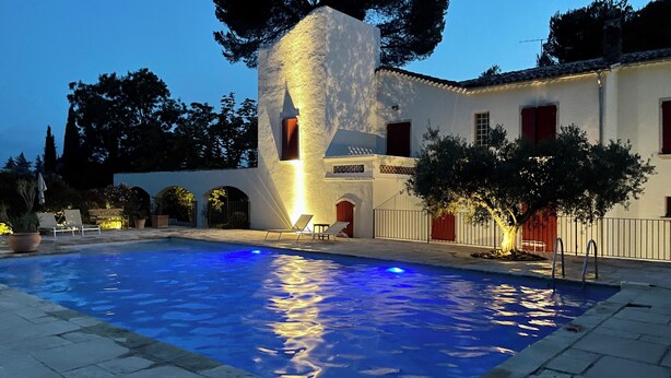 A pool area with a white building, red shutters, and a clear blue sky. - Large villa surrounded by pine trees in Provence (Sernhac)