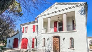 A two-story building with a white facade, red shutters, and a balcony with a railing. - Large villa surrounded by pine trees in Provence (Sernhac)