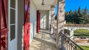 Un couloir extérieur abrité, doté de volets rouges, d’un ventilateur de plafond et offrant une vue sur un jardin.