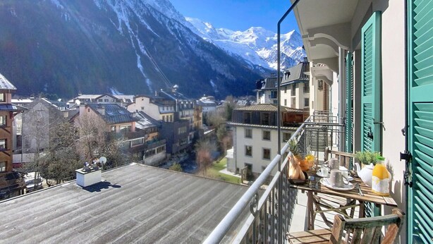 A balcony with a view of snow-capped mountains, a wooden table with breakfast items, and traditional architecture.