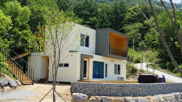 A modern two-story house with a stone retaining wall, a wooden staircase, and a view of a forested hillside. - Moonlight Village Pension (Danyang)