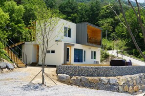 A modern two-story house with a stone retaining wall, a wooden staircase, and a view of a forested hillside. - Moonlight Village Pension (Danyang)