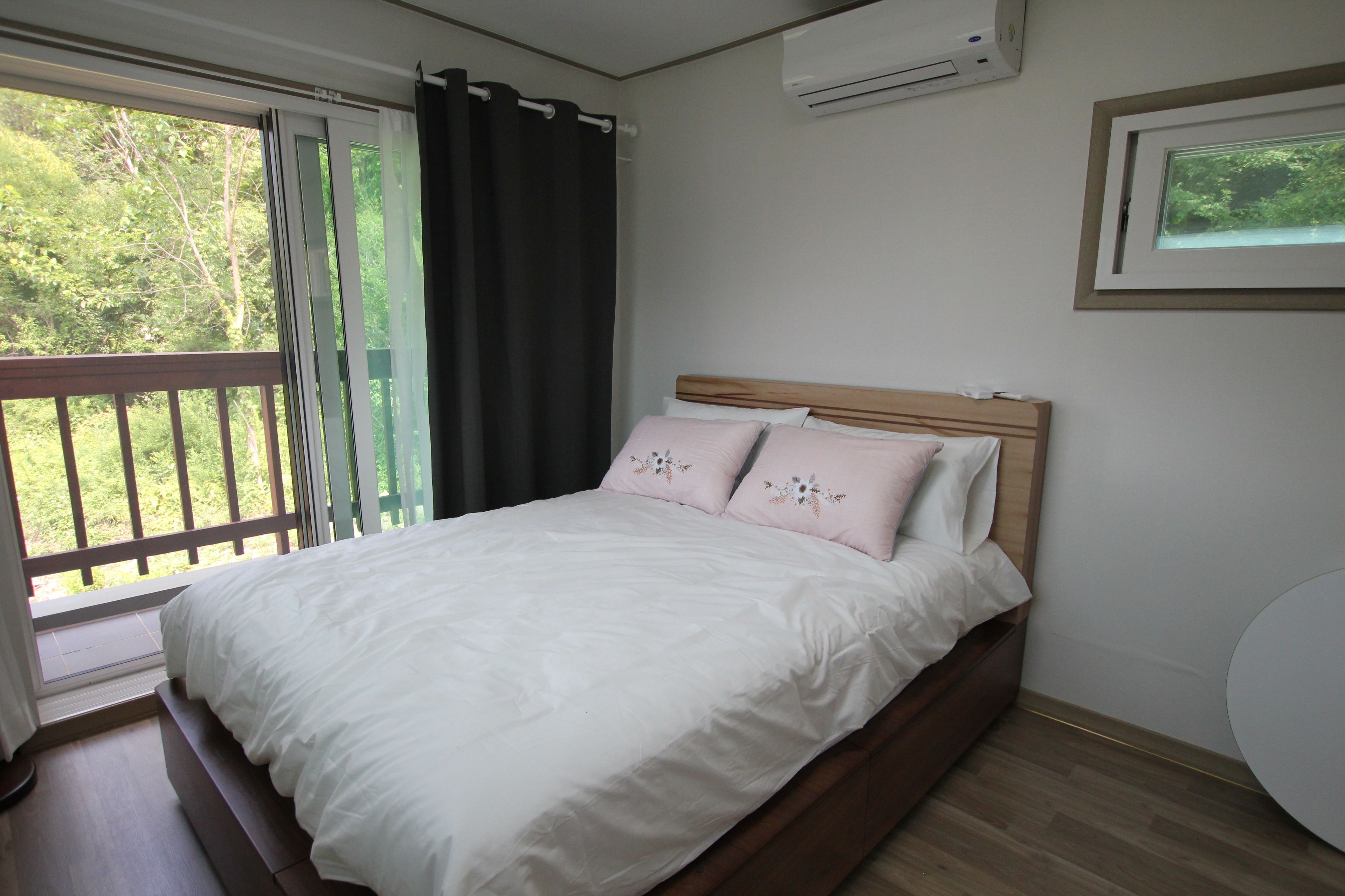 A bedroom with a bed, a window with curtains, an air conditioning unit, and a framed picture on the wall.