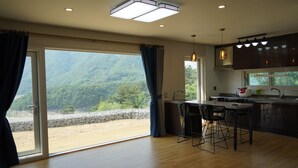 A modern kitchen with a large island, bar stools, and a view of a forested area through the sliding glass doors. - Moonlight Village Pension (Danyang)