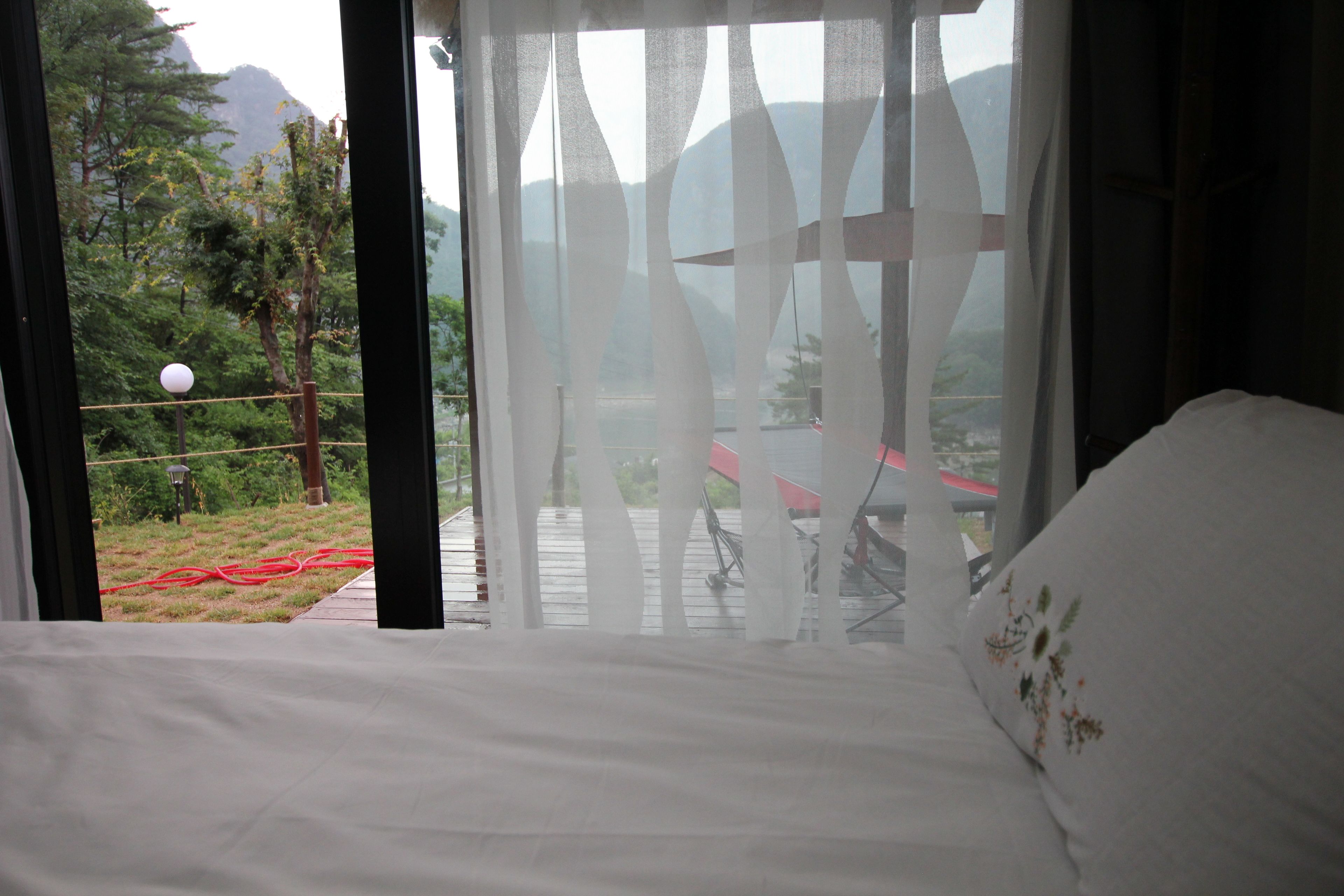 A bedroom with a bed, a view of a mountainous landscape through sheer curtains, and a red chair outside.