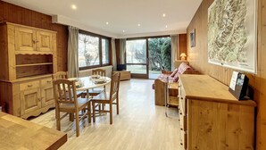 A spacious room with a wooden dining table, chairs, a sofa, and a large window offering a view of the outdoors. - Cozy Apartment in Chamonix, 4 Pers, Near Brévent with Terrace and Parking (Chamonix-Mont-Blanc)