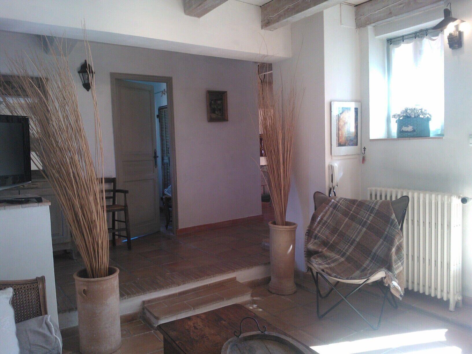 A living room with a fireplace, a television, and a chair with a plaid blanket. There are two tall vases with dried plants and a door leading to another room.