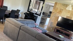 Una sala de estar con un sofá modular, una mesa de comedor con sillas, una zona de cocina y un televisor de pantalla plana sobre un soporte.