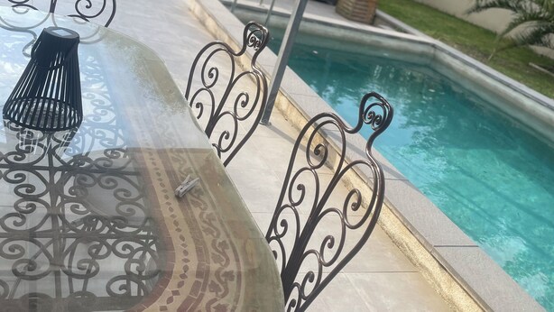 Un área junto a la piscina con un espacio de estar techado, una mesa de metal con diseños elaborados y una vista despejada de la pileta.