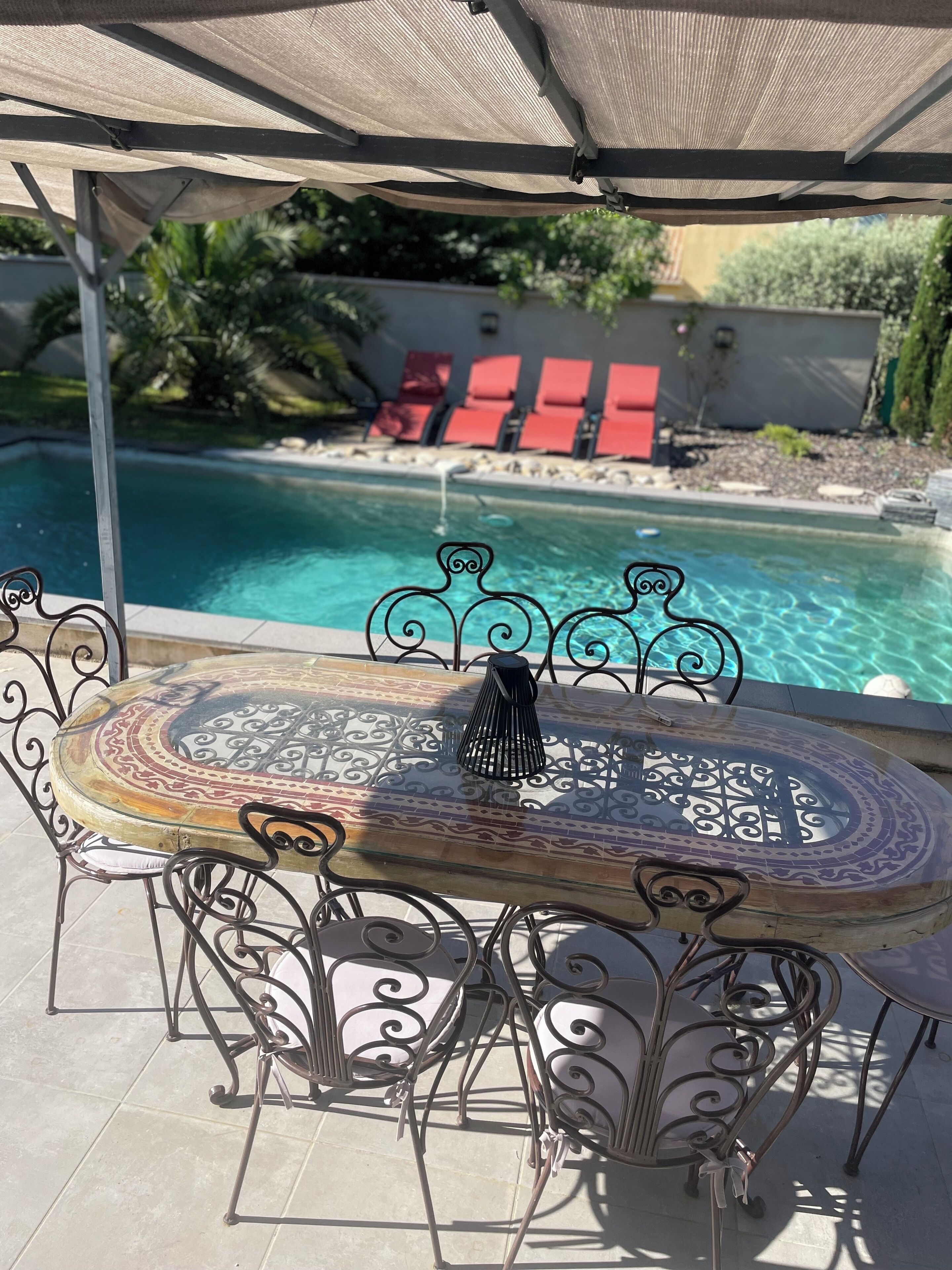 Una mesa junto a la piscina con tablero estampado y sillas de metal, ubicada en una zona sombreada con vista a una piscina y sillas de descanso rojas al fondo.