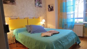 A bed with a green bedspread, a pillow, and folded towels. There is a lamp on a bedside table, a window with blue curtains, and a radiator. - CHAMBRE D'HOTE MERLOT CHEZ LE VIGNERON (LAURE MINERVOIS)