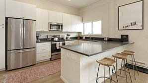 A modern kitchen with white cabinets, stainless steel appliances, a large island with bar stools, and a window with blinds. - 15 Chambertin Townhomes  3 Bedroom Townhouse by RedAwning (Avon)