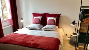A neatly made bed with a red blanket, white pillows, and a red and white pillow arrangement. There is a bedside lamp, a small round table with a lamp, and a woven basket on the floor. - Le Central, nice and quiet apartment in the center of Nancy (Nancy)