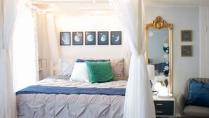 A bedroom with a canopy bed, a bedside table with a lamp, a mirror, and framed pictures on the wall.