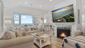 A spacious living room with a fireplace, a large TV mounted on the wall, and a view of the sea through large windows.