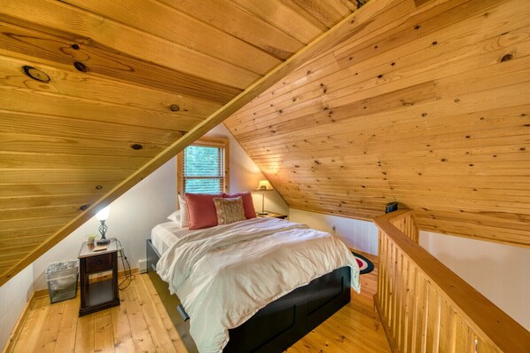 A bedroom with a sloped ceiling, wooden beams, a bed with pillows, a nightstand, and a window with blinds.