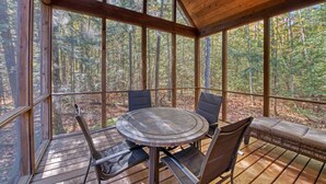Una terraza de madera con una mesa redonda y sillas, rodeada de amplios ventanales que ofrecen vistas a un bosque.