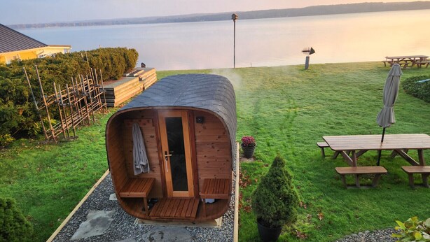 Un sauna en bois, doté d’une porte et d’un banc à l’intérieur, est installé sur une pelouse, à proximité d’une table de pique-nique et d’un parasol.