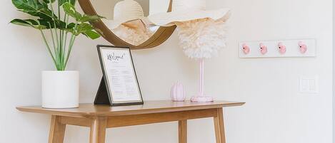 Een houten bureau met een spiegel, een lijst met een menukaart, een kamerplant en een roze lamp tegen een witte muur.