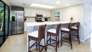 A modern kitchen with white cabinetry, a stainless steel refrigerator, a microwave, and a built-in oven. There are three wicker bar stools with white cushions.