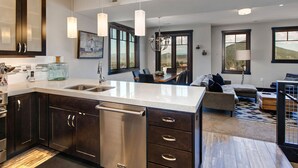 A modern kitchen with dark cabinetry, a dishwasher, and a large island with a sink. There is a dining area with a table and chairs, and a sofa in the background. - Luxe Townhome in Prime Location, Private Hot Tub, Chef (Park City)