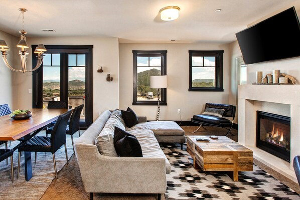 Una sala de estar moderna con chimenea, un comedor con mesa y sillas de madera, y ventanas amplias que ofrecen vistas a la montaña.