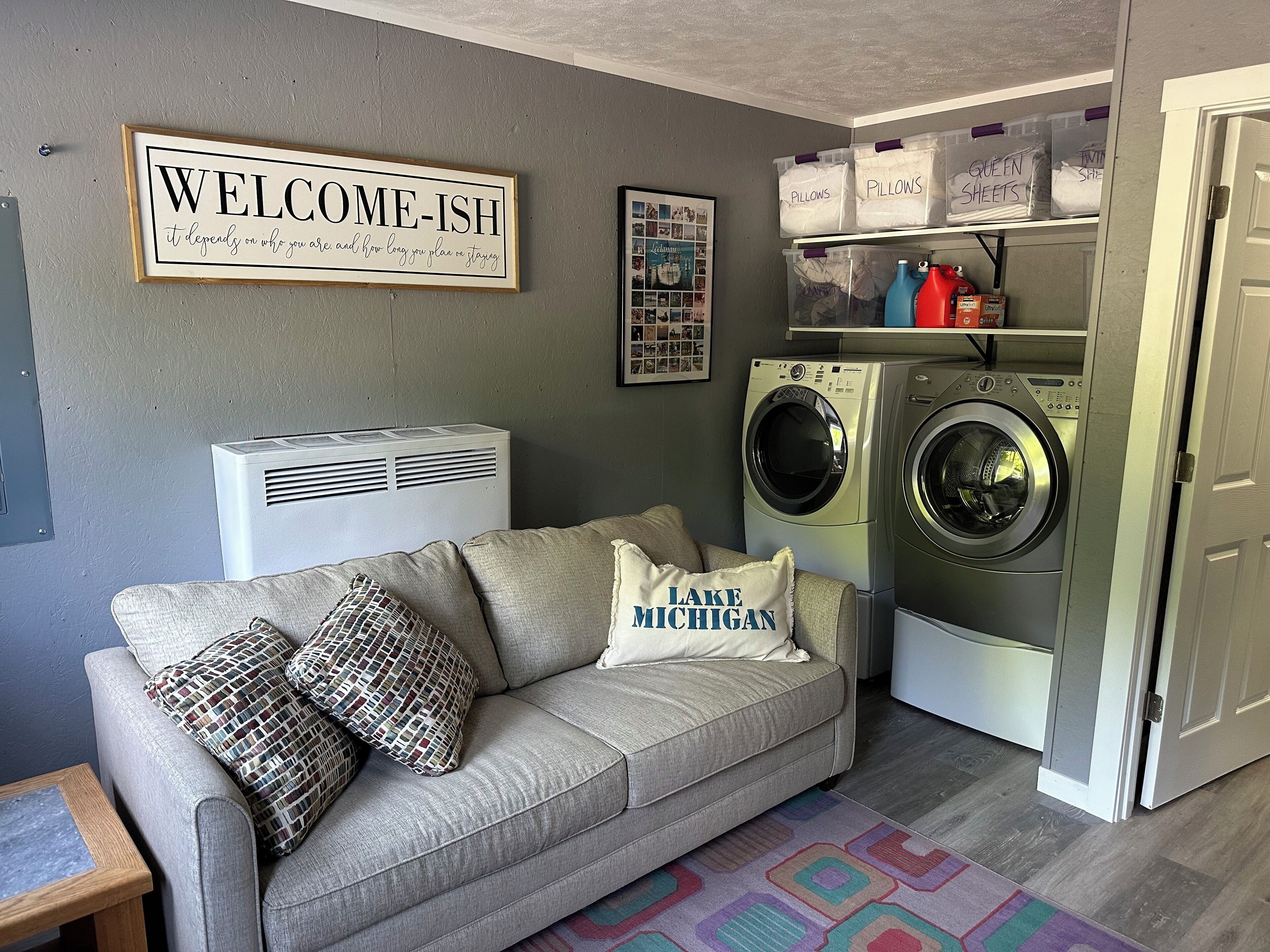 A laundry room with a gray couch, two washing machines, and storage bins labeled 'Pillows' and 'Queen Sheets'.