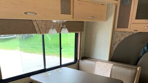A compact RV interior with a kitchenette, a dining area, and a window providing a view of the outdoors.