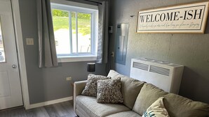A room with a grey couch, patterned pillows, a colorful rug, a window with curtains, and a wall sign saying "WELCOME-ISH".