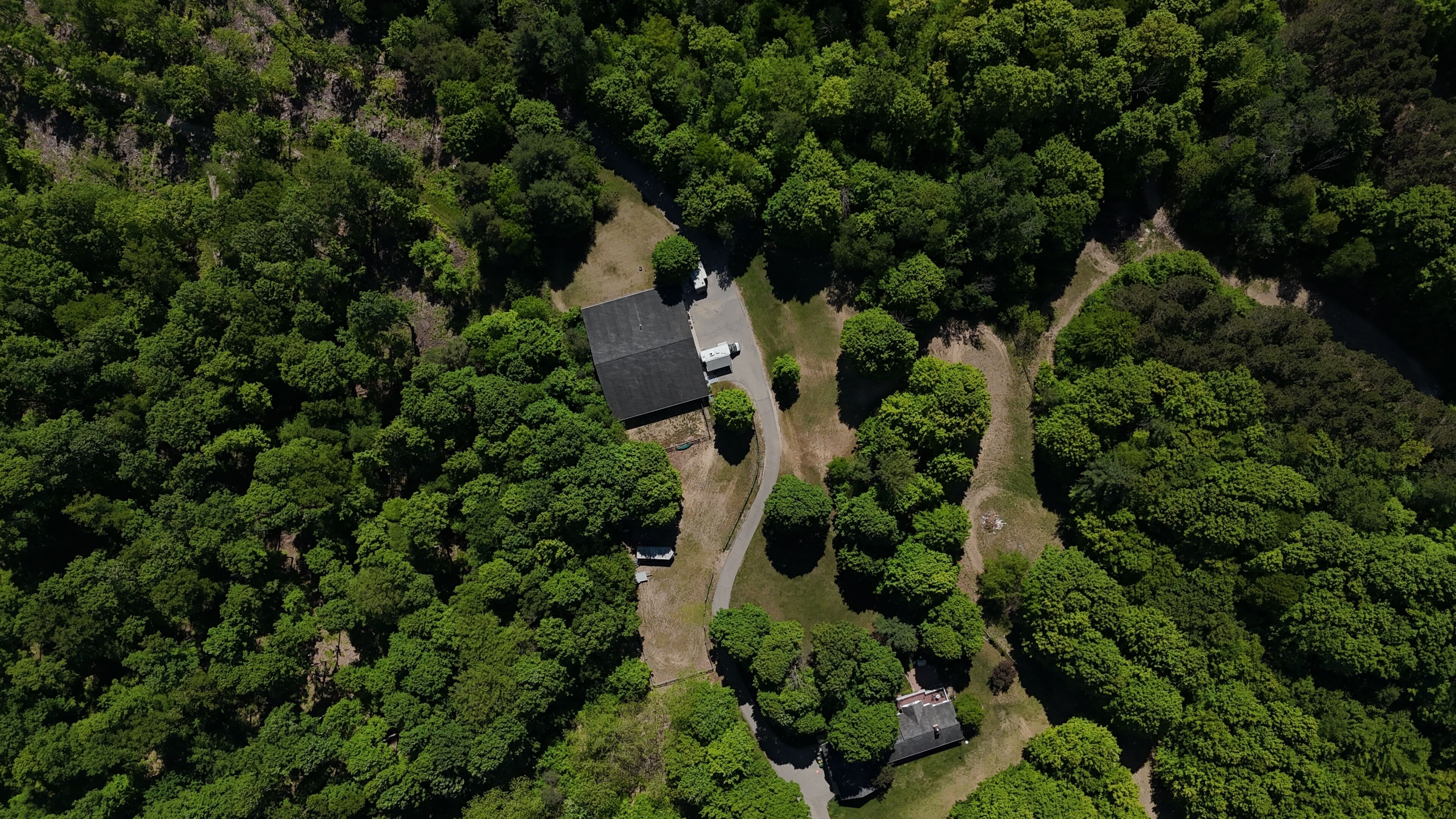 An aerial view of a property surrounded by dense forest, featuring a large building with a flat roof and a smaller structure nearby.
