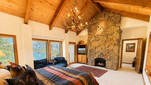 Ein Schlafzimmer mit einem großen Bett, einem Kamin aus Stein, einer Chandelüster und Blick ins Freie.