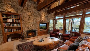 Ein gemütliches Wohnzimmer mit Kamin aus Stein, Bücherregalen, Ledersofas und großen Fenstern mit Blick auf die Berge.