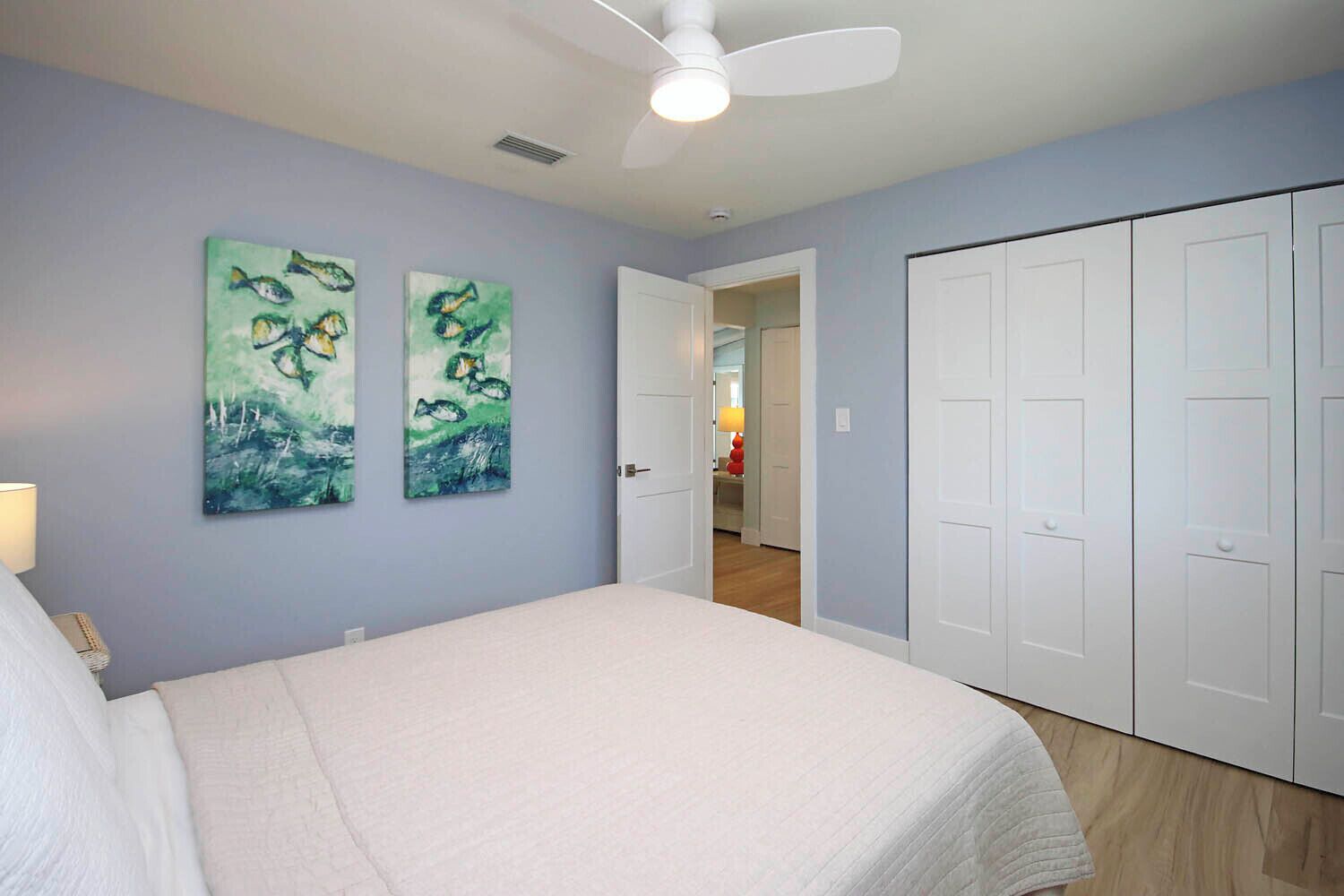 A bedroom with a bed, two paintings, a ceiling fan, and a set of white doors.
