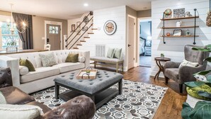 Un salon moderne comprenant un canapé, un pouf et des fauteuils, un escalier et un couloir menant à une chambre.
