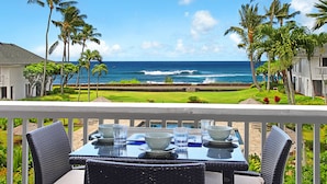 Una mesa de comedor con sillas en un balcón con vistas a una playa con palmeras y olas del océano.