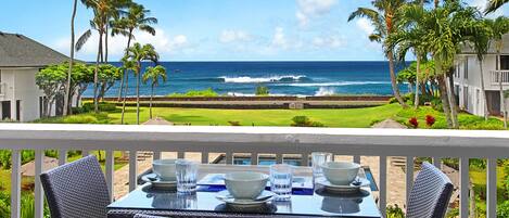 A dining table with chairs on a balcony overlooking a beach with palm trees and ocean waves.