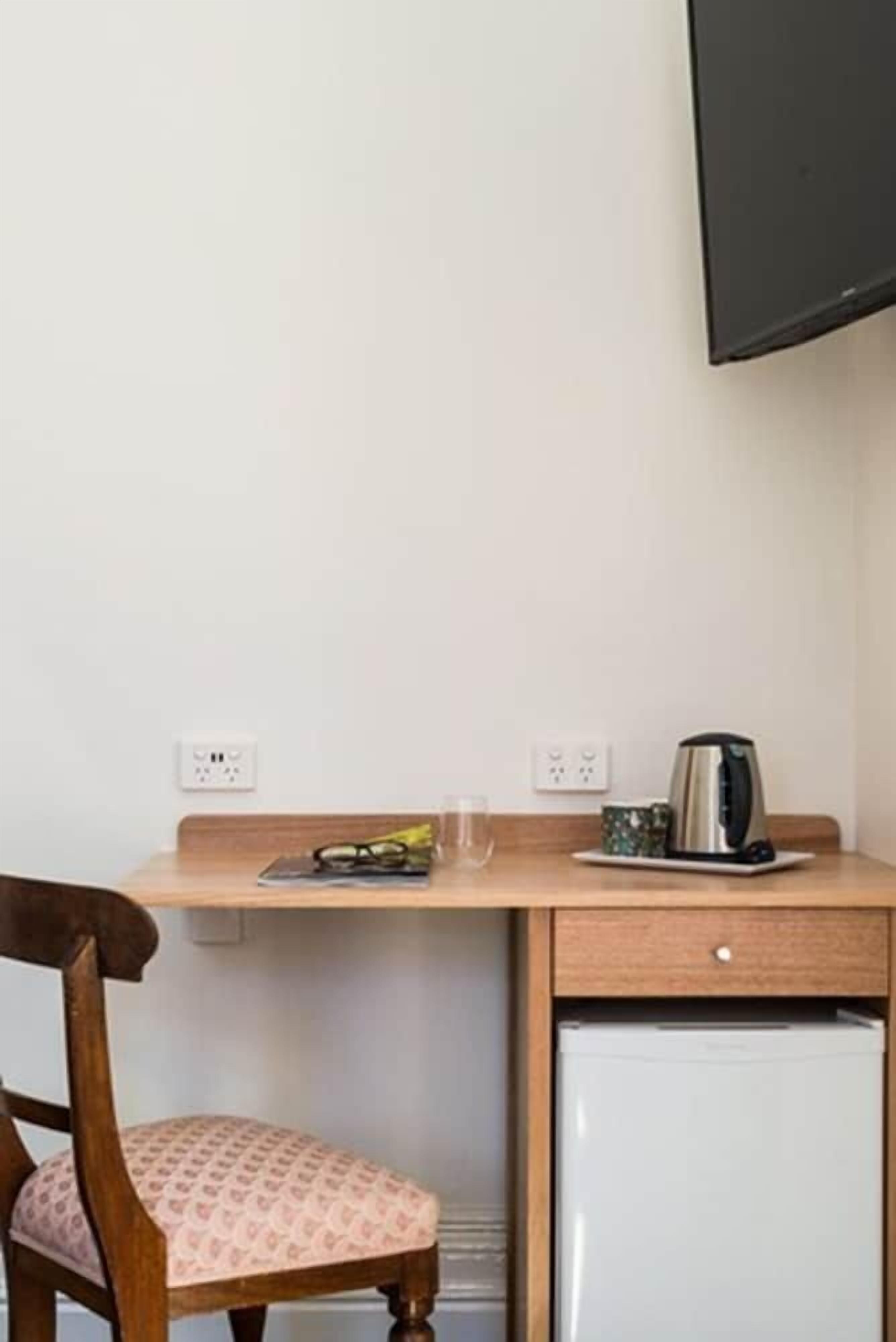 A compact room with a wooden desk, a chair, a small refrigerator, a kettle, and a flat-screen TV mounted on the wall.