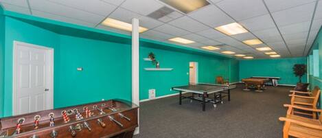 A room with teal walls, a foosball table, a ping pong table, and a pool table.