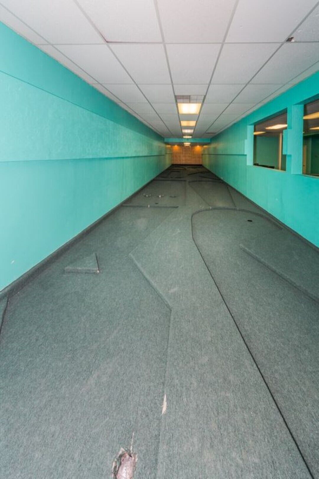 A corridor with teal walls and a grey floor, leading to a room with a window.