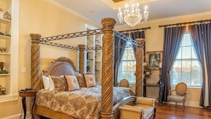 Ein Bett mit Baldachin, reich verziert mit Schnitzereien, eine Kristallleuchter und ein klassischer Sessel in einem wohlig beleuchteten Raum mit Holzboden.