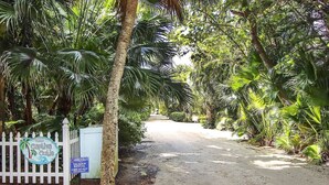 A dirt road flanked by lush tropical vegetation, leading to a white picket fence with a sign indicating a vacation rental.